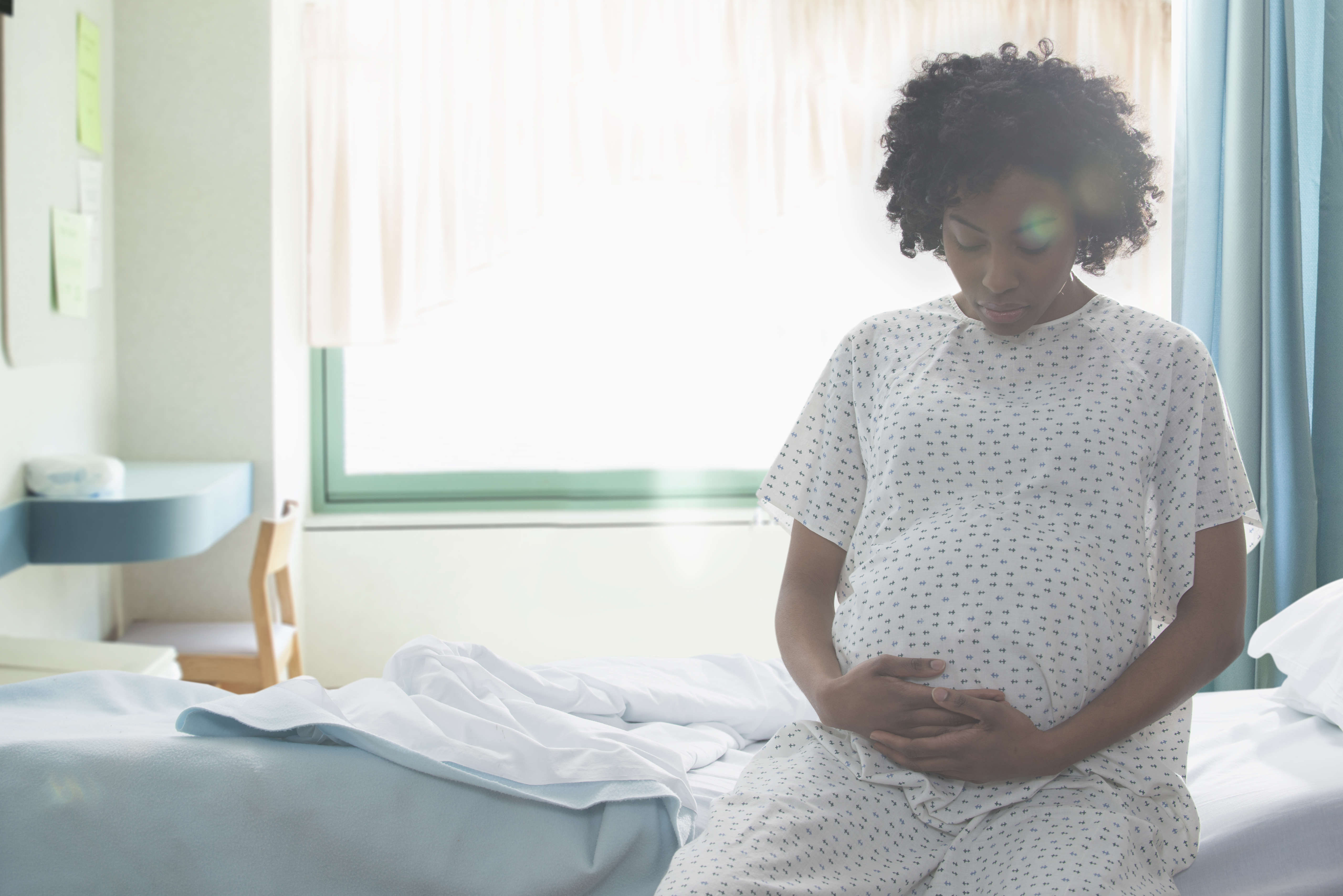 Pregnant woman sitting down on hospital bed with white gown on.