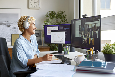Woman smiling and looking at PC monitors with headphones on sitting down.